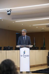 Um homem pardo de cabelos grisalhos fala atrás de um púlpito, atrás, um auditório moderno com bandeiras, mesas e computadores