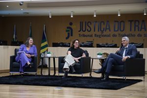 Duas mulheres e 1 homem estão sentados em poltronas em cima de um palco do auditório, um deles fala ao microfone e os demais olham para ele
