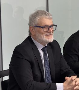 Uma foto em close-up de um homem com cabelos e barba grisalhos, usando óculos de armação azul e um terno escuro com uma gravata estampada. Ele está sorrindo e olhando para o lado direito da imagem.