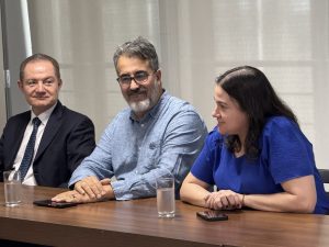 Uma mulher e dois homens sentados em uma mesa de madeira. O homem da esquerda veste terno e gravata e olha para frente com um leve sorriso. O homem do meio tem barba e usa óculos e uma camisa azul clara, sorrindo e olhando para a mulher à sua direita. A mulher, também sorrindo, usa uma blusa azul escura e está com as mãos sobre a mesa, olhando para o homem do meio.