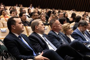 A imagem mostra um auditório com várias pessoas sentadas em poltronas claras, algumas delas reservadas.
