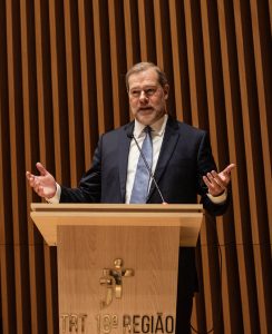 Homem de terno em um púlpito com microfones, gesticulando. Ao fundo, parede de madeira. No púlpito, um logotipo com "TRT 18ª REGIÃO".