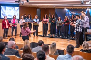 Um grupo de pessoas, a maioria mulheres, canta em um palco em frente a uma plateia. Há um telão ao fundo mostrando "PRÊMIO TRT".







