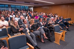 A imagem mostra uma plateia sentada em um auditório, com cadeiras azuis estofadas e encosto de madeira. A maioria das pessoas está aplaudindo, com as mãos na altura do peito. Há homens e mulheres de diversas idades, vestidos com trajes formais e semi-formais. O auditório tem paredes de madeira e iluminação embutida no teto. As primeiras filas estão mais em foco, enquanto as filas de trás estão um pouco desfocadas.