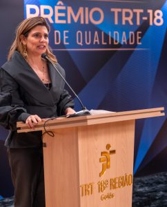 A imagem mostra uma mulher de meia-idade, com cabelo castanho-claro e brincos grandes, falando em um púlpito de madeira. Ela veste um casaco escuro com gola volumosa e calças escuras. Ao fundo, um painel azul com o texto "PRÊMIO TRT-18 DE QUALIDADE". No púlpito, há o logotipo e o texto "TRT 18ª REGIÃO Goiás". A iluminação é focada na mulher e no púlpito.