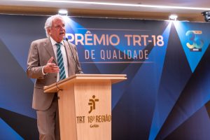 A imagem retrata um evento do Tribunal Regional do Trabalho da 18ª Região (TRT-18), localizado em Goiás, especificamente a cerimônia do "Prêmio TRT-18 de Qualidade". Um homem de terno está discursando em um púlpito, com um painel ao fundo indicando a celebração dos "33 anos" do TRT-18.







