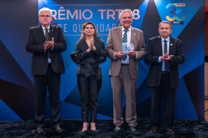 Quatro pessoas, sendo três homens e uma mulher, em pé, em um palco com iluminação azul, e o texto "PRÊMIO TRT18 DE QUALIDADE". O homem à direita do centro segura um troféu.