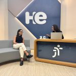 foto de longe mostra a entrevistadora sentada atrás de uma bancada, olhando para a entrevistada, que está sentada de pernas cruzadas em um sofá cinza e olhando para a entrevistadora. Atrás delas um painel azul e cinza com a logo do programa.