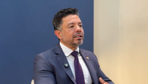 Um homem pardo de cabelos escuros e barba grisalha está de terno, um pouco de lado, sentado falando