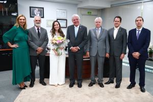 A foto mostra um grupo de sete pessoas, cinco homens e duas mulheres, posando formalmente em um escritório. A mulher no centro da imagem está segurando um buquê de flores. Todos estão vestidos com trajes sociais, a maioria ternos, e a mulher à esquerda usa um vestido verde. O ambiente é bem iluminado e parece ser um escritório ou sala de reunião, com quadros e diplomas nas paredes.