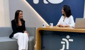Uma mulher sentada em uma poltrona e um mulher numa bancada. Elas olham para a câmara. Ao fundo, a logomarca do programa hora extra. Embaixo da bancada, a logomarca da Justiça do Trabalho