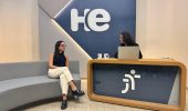 Uma mulher sentada em uma poltrona e um mulher numa bancada. Elas olham para a câmara. Ao fundo, a logomarca do programa hora extra. Embaixo da bancada, a logomarca da Justiça do Trabalho