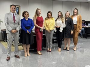 A imagem mostra sete pessoas, um homem e seis mulheres, posando em um escritório. O homem veste um terno cinza com gravata bordô. As mulheres vestem roupas variadas: uma blusa azul com saia preta, um terno vermelho, um blazer amarelo com calças cinzas, uma blusa de renda branca com calças pretas, um blazer preto com calças pretas, e um terno cor de camelo.