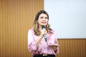 A imagem mostra uma mulher com cabelos castanhos claros, vestindo uma camisa de seda rosa e calças escuras, falando em um microfone e segurando um celular rosa. Ela está em um ambiente com parede de madeira clara e um quadro branco ao fundo.