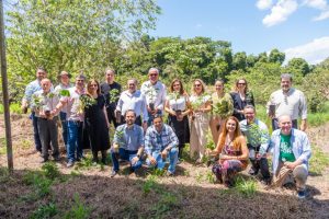 A imagem mostra um grupo de cerca de 20 pessoas, em sua maioria adultos, posando ao ar livre em uma área com vegetação.Algumas pessoas estão de pé na parte de trás, enquanto outras estão agachadas na frente. Quase todas as pessoas seguram mudas de árvores em vasos pequenos, indicando que participam de uma ação de plantio.O ambiente é rural ou de área verde, com árvores e arbustos visíveis no fundo. O tempo parece ensolarado.
