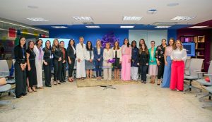 Um grupo de aproximadamente quinze mulheres bem-vestidas está posando para uma foto em um escritório moderno com paredes coloridas e cadeiras de escritório.