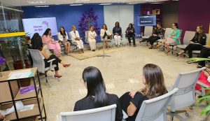 Um grupo de mulheres está sentado em um círculo em um ambiente de escritório moderno, participando de um evento. Telas ao fundo exibem "Flores do Ipê" e "Reimagine Carr...", e uma árvore decorativa roxa se destaca contra a parede azul.