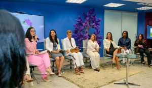 Seis mulheres estão sentadas em cadeiras dispostas em semicírculo em um ambiente de escritório moderno, enquanto uma sétima mulher, vista parcialmente à esquerda, parece estar falando. Ao fundo, uma grande tela exibe uma ilustração floral e uma árvore decorativa roxa se destaca contra a parede azul.