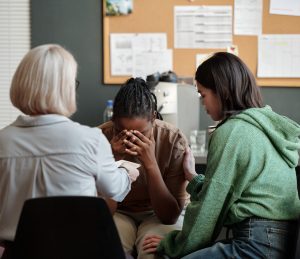 Uma mulher com as mãos no rosto, aparentemente em sofrimento, está sentada entre duas outras mulheres, uma delas com a mão no ombro da mulher aflita, e a outra, de costas, estendendo um lenço. O fundo mostra um mural de cortiça com papéis.