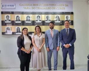 A imagem mostra um grupo de quatro pessoas, dois homens e duas mulheres, em um ambiente interno. Eles estão posando de pé, sorrindo e olhando para a câmera. Ao fundo, há uma parede com a inscrição "GALERIA DOS JUIZES TITULARES DA VARA DO TRABALHO DE CERES" e uma série de fotos de juízes. Há também a bandeira do Brasil à esquerda e, na parte superior, o logotipo "TRT - 18ª REGIÃO Goiás".