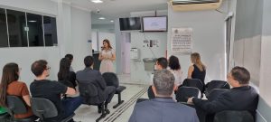 A imagem mostra uma mulher de pé, vestindo um vestido bege, apresentando algo para um grupo de pessoas sentadas em cadeiras de escritório, em um ambiente interno. A mulher aponta para uma tela de TV na parede, onde se lê "Saúde e autogestão". Há um bebedouro e uma placa com um texto longo também na parede. O público, composto por homens e mulheres, está de costas para a câmera, com exceção de alguns.
