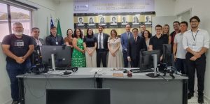 A imagem mostra um grupo de cerca de 15 pessoas, homens e mulheres, de pé atrás de uma mesa de escritório com computadores. Ao fundo, uma galeria de fotos na parede exibe a inscrição "GALERIA DOS JUIZES TITULARES DA VARA DO TRABALHO DE CERES" e fotos em preto e branco. O grupo posa para a foto.