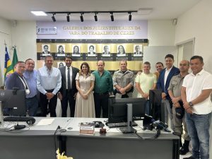 A imagem mostra um grupo de cerca de 15 pessoas, homens e mulheres, posando para uma foto em um escritório. Todos estão de pé e a maioria está vestindo trajes sociais ou casuais. Ao fundo, uma parede exibe uma galeria de fotos de juízes com a inscrição "GALERIA DOS JUIZES TITULARES DA VARA DO TRABALHO DE CERES". A foto foi tirada por trás de uma grande mesa com computadores e documentos.