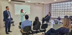 Uma mulher está de pé, apresentando uma palestra para um grupo de pessoas sentadas. Ao lado dela, um homem de terno a observa. Na tela de projeção está escrito "Saúde e Qualidade".