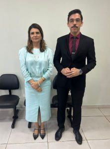 Uma mulher e um homem estão de pé, lado a lado, posando para a foto. Ambos estão vestidos socialmente.