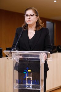 Uma mulher de óculos está em pé em um púlpito de acrílico, falando em um evento. No púlpito, há o logo do TRT-18ª Região.