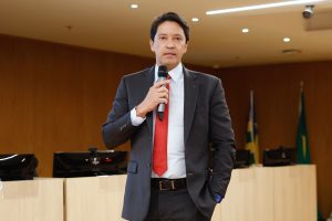  Um homem de terno e gravata vermelha está em pé no palco, segurando um microfone, falando para a plateia.