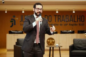 Um homem de terno, barba e óculos está em pé, no palco, segurando um microfone e um controle, enquanto fala para a plateia.