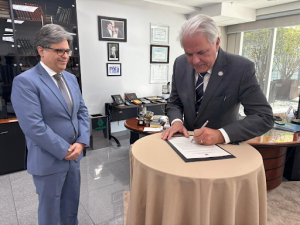 Dois homens em pé na foto. Um deles assina um documento enquanto o outro observa. Eles usam terno.