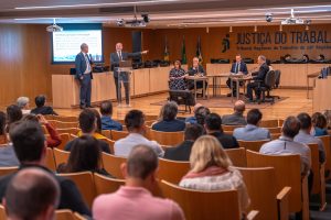 Foto do auditório do TRT GO com o público de costas e os palestrantes no palco. Do lado esquerdo 2 homens em pé, e no centro 5 pessoas simulam uma audiência de conciliação.