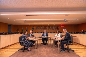 Grupo de pessoas estão ao redor de uma mesa no centro do palco e simulam uma audiência de conciliação