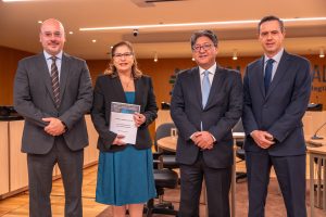 Foto com 4 pessoas no palco, 3 homens e 1 mulher, todos estão de roupas sociais, a mulher segura um relatório nas mãos.