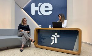 Uma mulher sentada em uma poltrona e um mulher numa bancada. Elas olham para a câmara. Ao fundo, a logomarca do programa hora extra. Embaixo da bancada, a logomarca da Justiça do Trabalho
