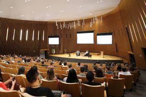 Uma visão ampla de um auditório moderno e lotado, com a plateia sentada em poltronas de madeira. No palco, há dois homens conversando em poltronas e três telões brancos ao fundo.