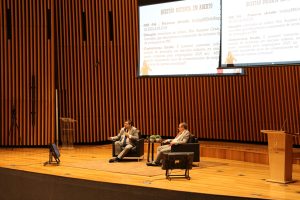 Dois homens estão sentados em poltronas no palco de um auditório, conversando. Ao fundo, dois telões exibem uma apresentação com o texto "QUESTÃO DISTINTA EM ABERTO". O auditório tem paredes de madeira.