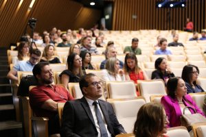 Uma vista parcial da plateia de um auditório, mostrando um grande número de pessoas sentadas em fileiras de poltronas bege, atentas a um evento ou apresentação.