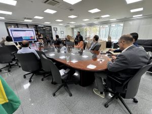 Várias pessoas sentadas numa mesa oval assistindo a uma apresentação