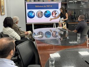 Pessoas sentadas em uma mesa oval assistem a uma apresentação de uma mulher que mostra um painel. Ela está em pé em frente à mesa.