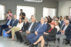 Várias pessoas sentadas em um auditório. Elas usam roupas formais