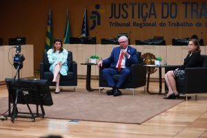 Três pessoas sentadas em um auditório com a logomarca da Justiça do Trabalho de Goiás ao fundo