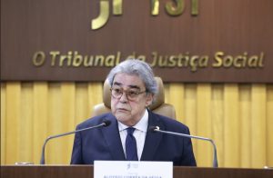 Homem de terno fala ao microfone. Ele está sentado