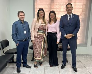 A imagem mostra um grupo de quatro pessoas - dois homens e duas mulheres - em pé, posando juntos em um ambiente interno. Todos estão sorrindo. O grupo está vestido com trajes de negócios ou social-casual. A foto parece ter sido tirada em um escritório ou sala de espera, com cadeiras e persianas visíveis ao fundo.