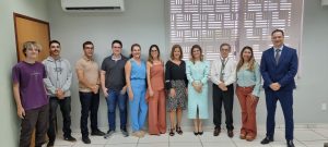 11 pessoas de pé, lado a lado, sorrindo e posando para foto. São cinco mulheres e seis homens. Ao fundo, uma parede cinza.