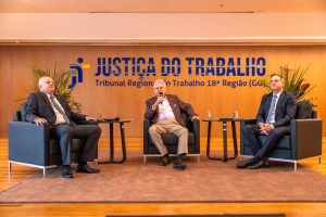 Três homens de terno estão sentados em poltronas pretas sobre um tapete no palco de um auditório. O homem do centro está segurando um microfone.