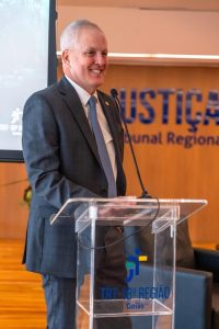 Um homem sorridente de terno está em pé atrás de um púlpito de acrílico em um palco.