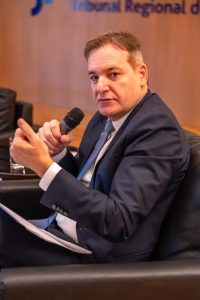 Um homem de terno está sentado em uma poltrona, segurando um microfone e gesticulando com a mão direita, enquanto olha para a câmera.